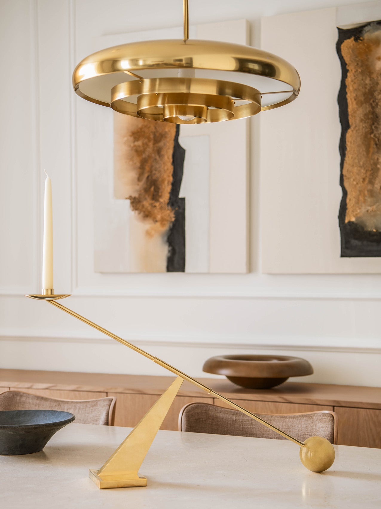 Brushed brass concentric-ring pendant light and matching angled brass candle holder on a neutral dining table