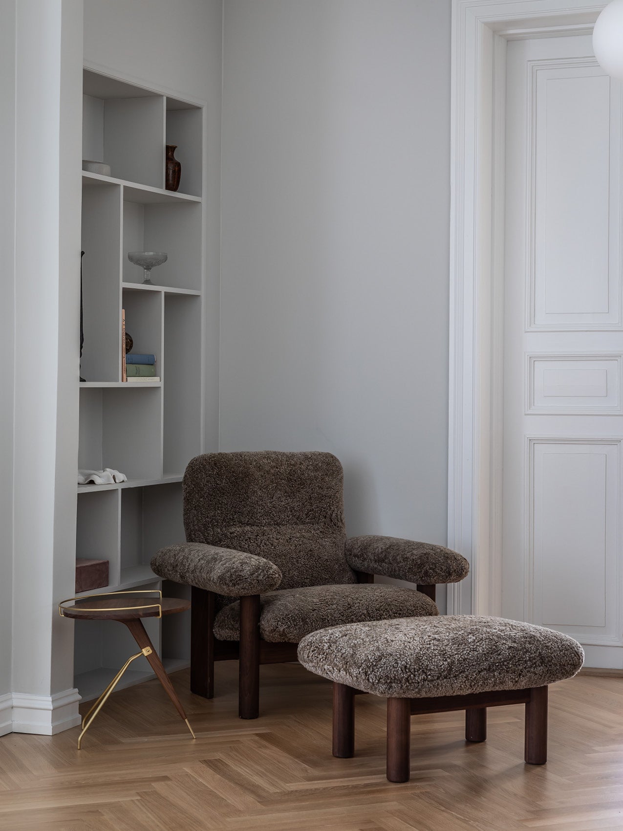 Brown sheepskin armchair with matching ottoman on dark wooden legs beside a small brass-and-wood side table and built-in