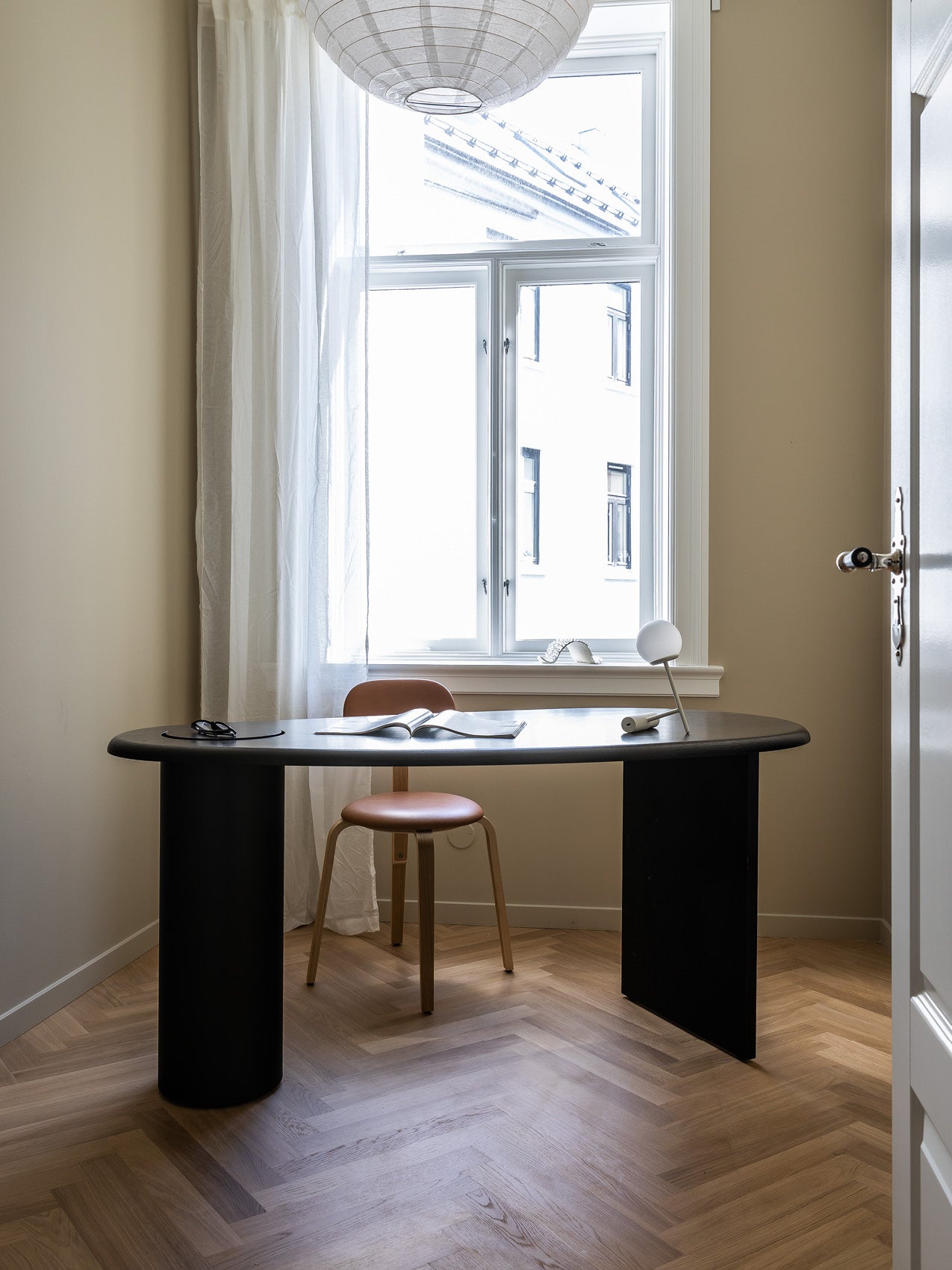 Minimal beige room with black oval desk, tan wooden stool, white desk lamp and sheer curtains by a bright window