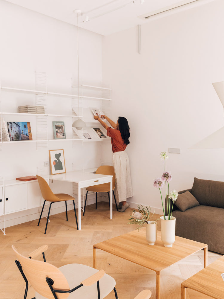 Minimal Scandinavian living room with light wood tables and chairs, white shelving, beige sofa and a person placing a book