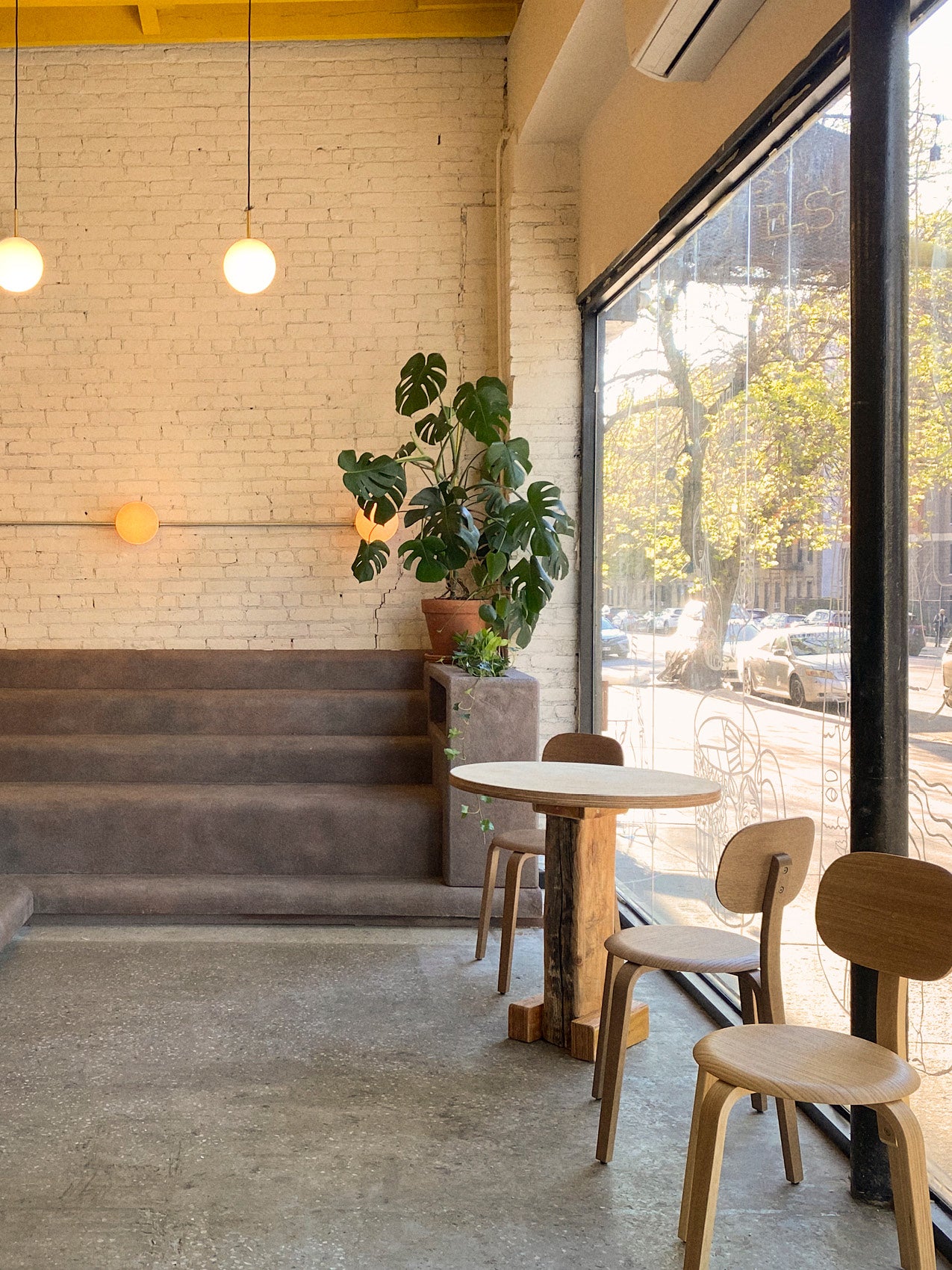 Minimal café interior with round wooden table and chairs, concrete steps, potted monstera, globe pendant lights, large window