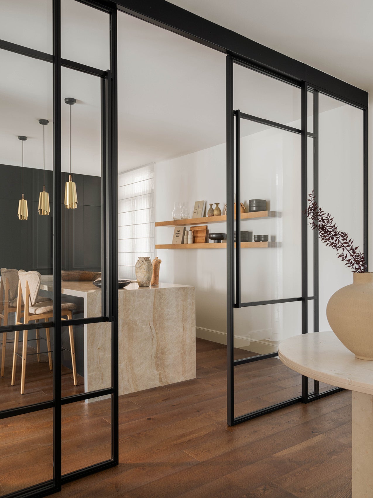 Black-framed glass sliding room divider revealing a neutral dining area with marble island, wooden shelves and gold pendant