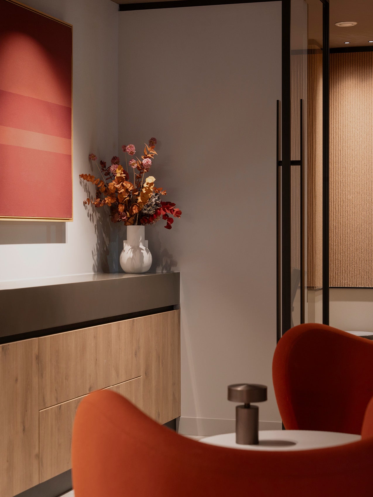 White ceramic vase with dried orange and red foliage on grey sideboard beside coral artwork, red lounge chair foreground
