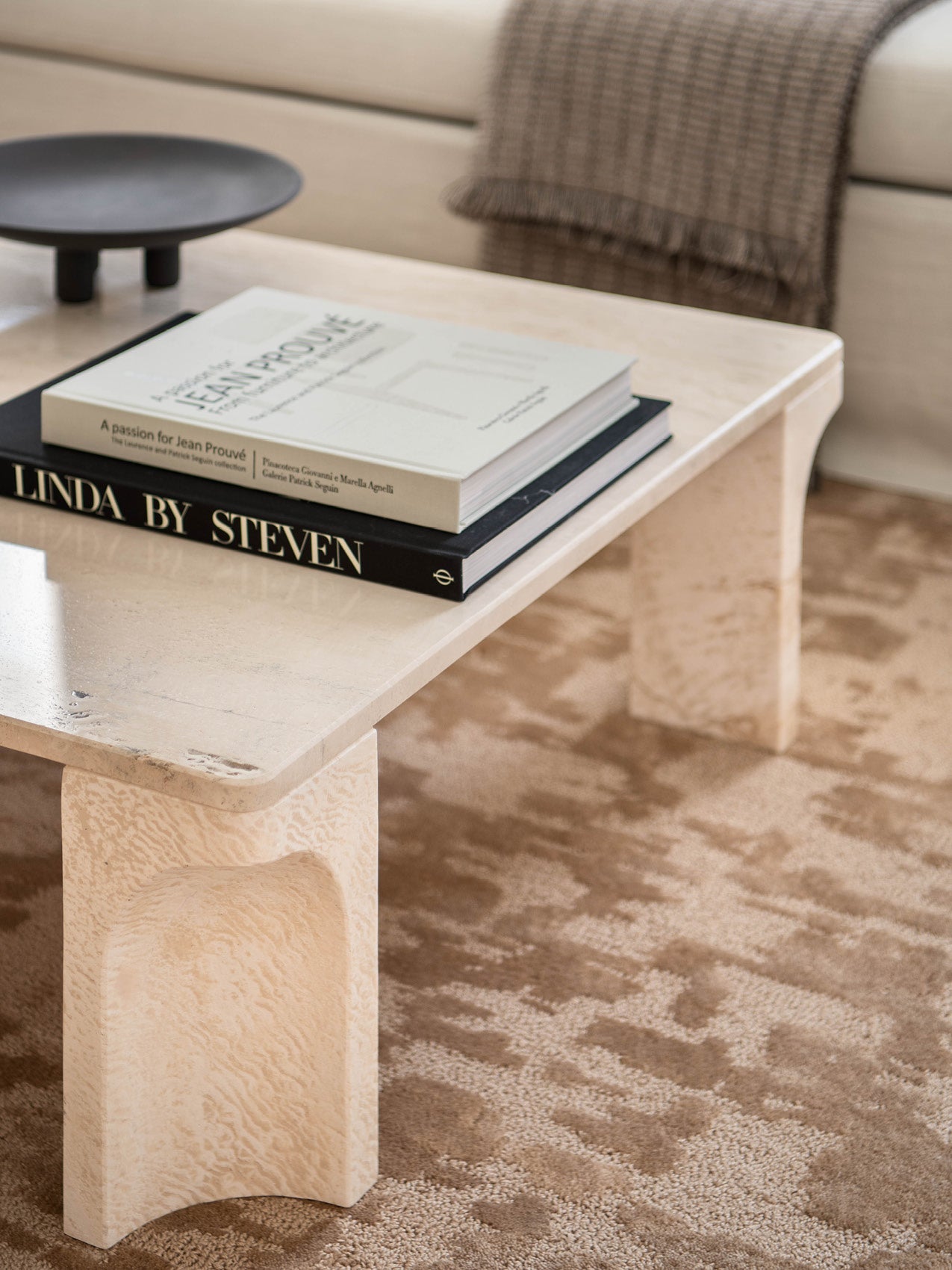 Beige travertine coffee table with sculpted legs, stacked black and white design books and a matte black round tray.