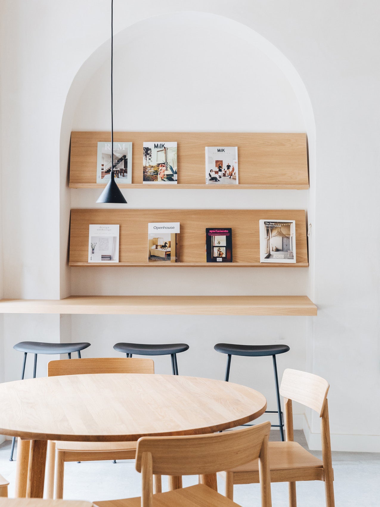 Minimal dining scene with round light wood table, chairs and stools; recessed wall shelves with oak magazine ledges and