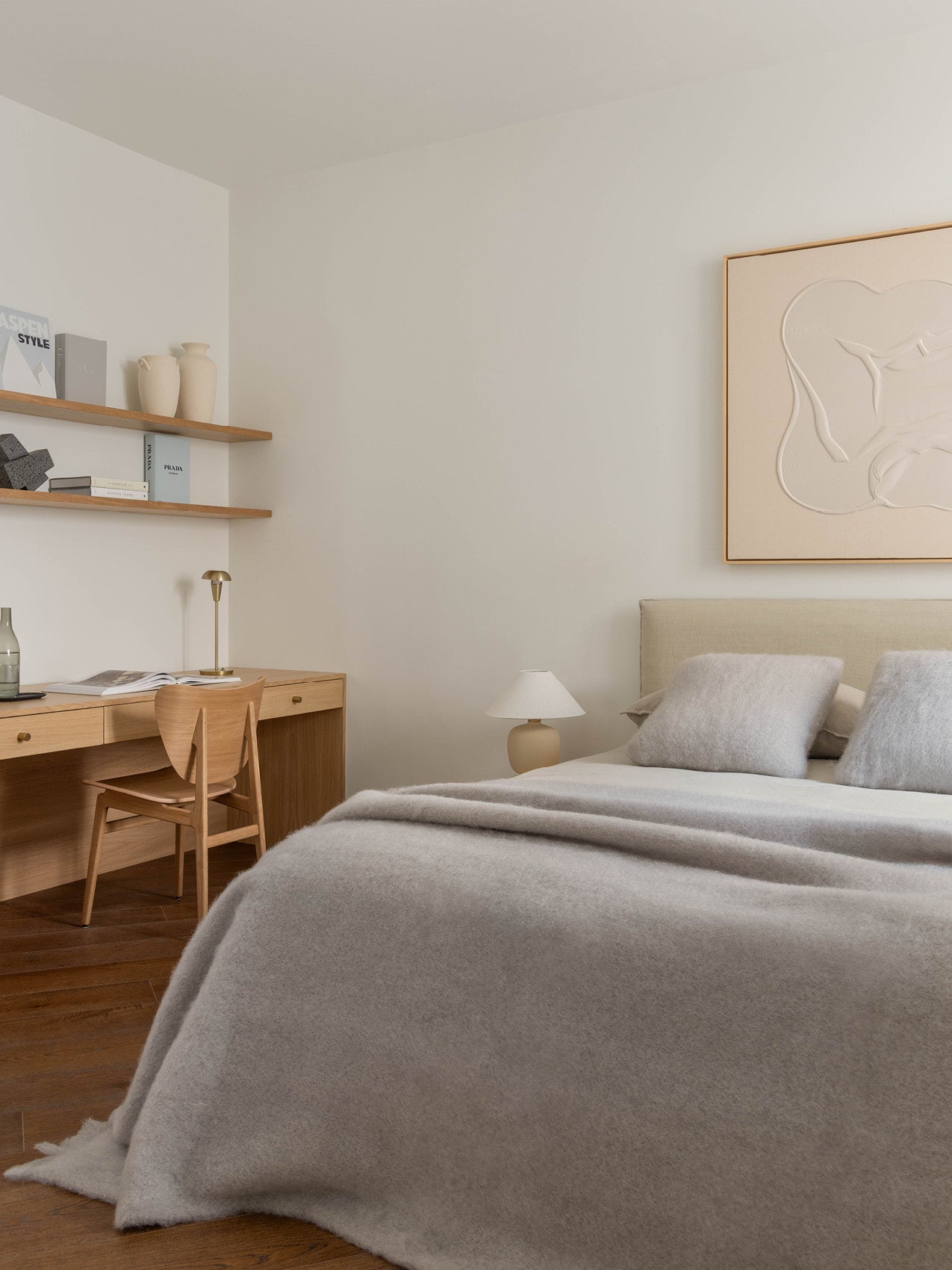 Minimal bedroom with wool grey bedspread and pillows, beige upholstered headboard, wooden desk and chair, ceramic lamp and