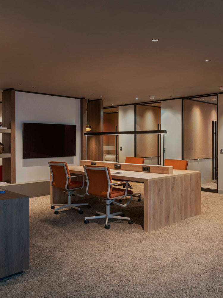 Modern co-working desk in light oak with integrated power, two orange upholstered swivel chairs and overhead black linear