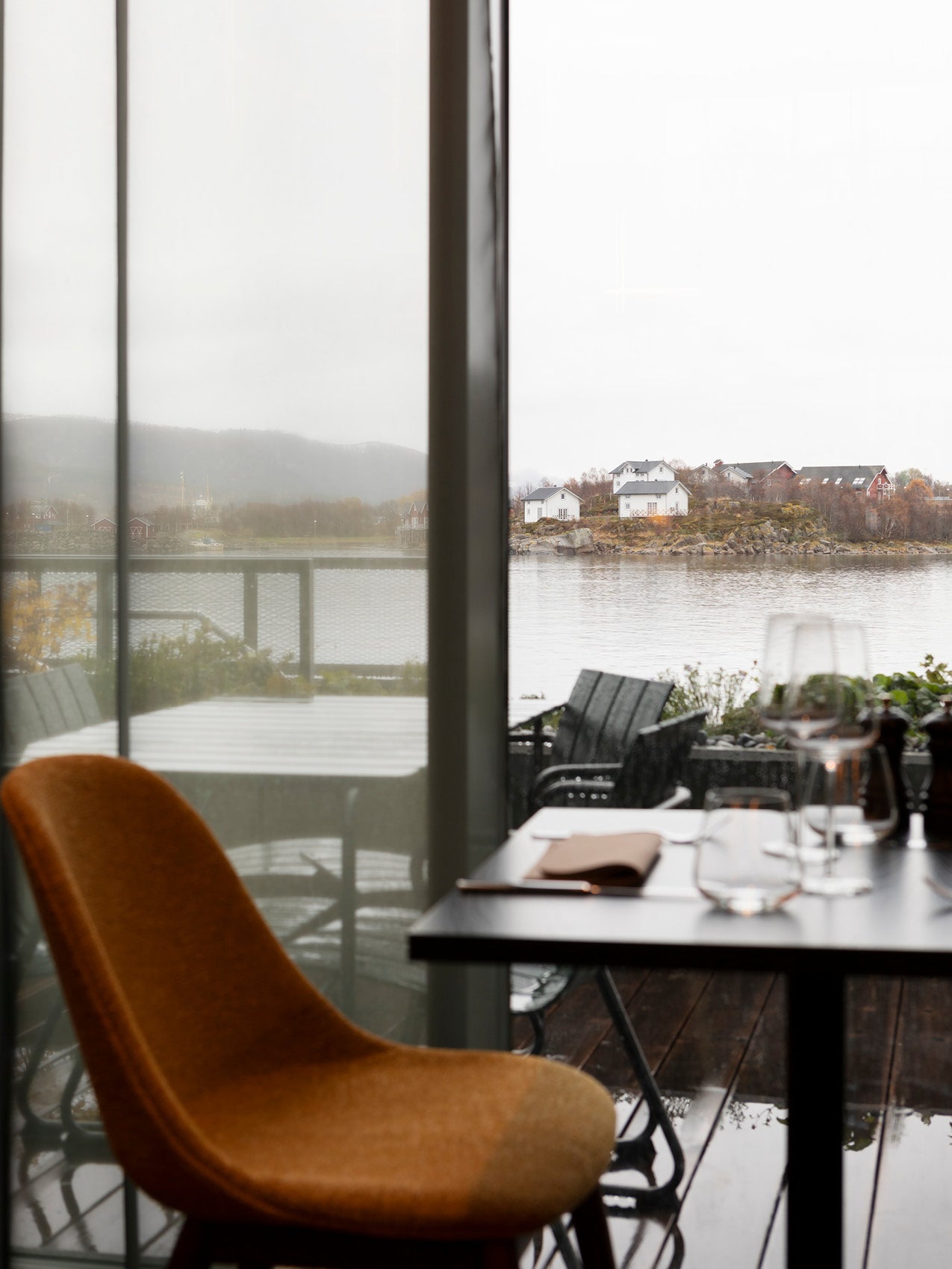 Rust-coloured upholstered curved dining chair by a glass table, overlooking a rainy balcony and white coastal houses across