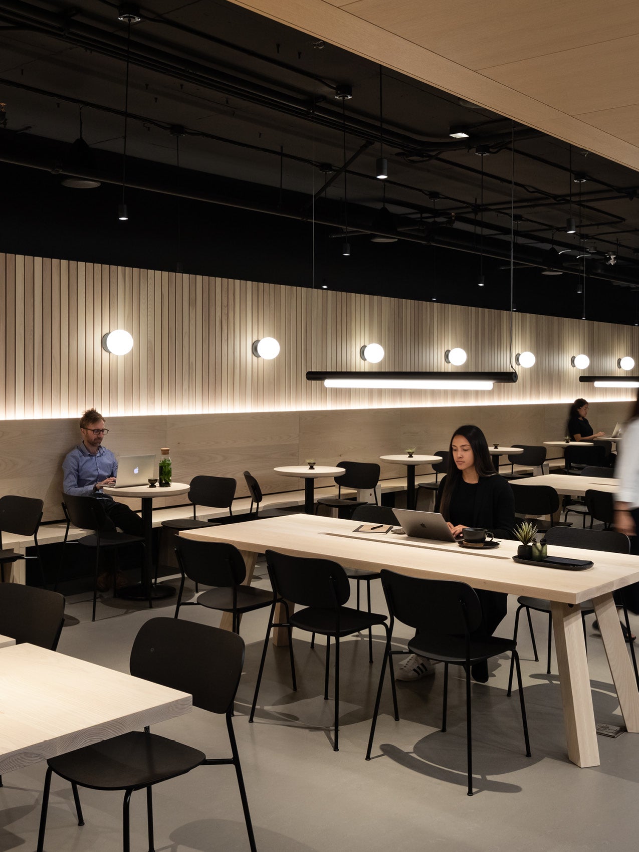 Minimalist cafe interior with light oak communal tables, black chairs, round wall lights and people working on laptops