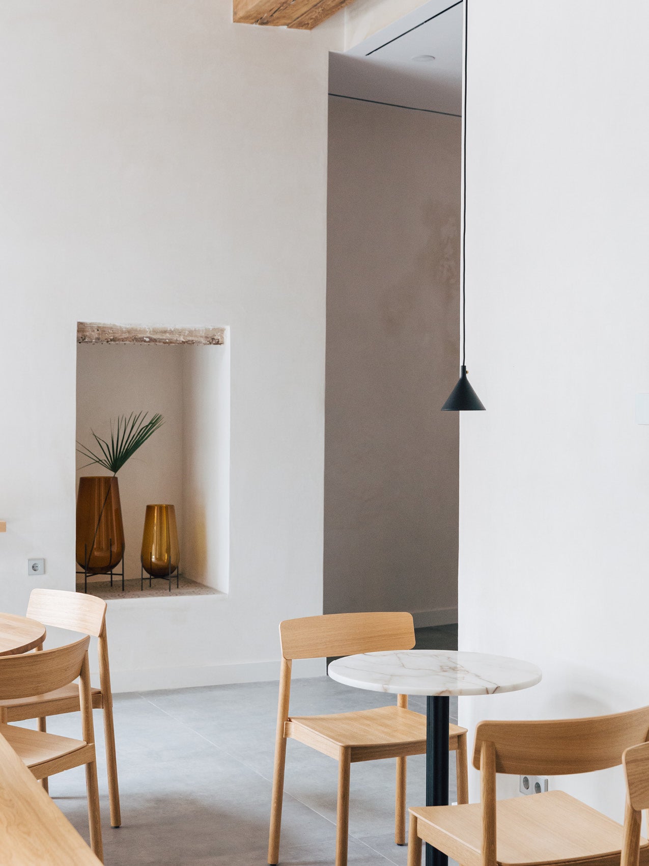 Minimal dining corner with light oak chairs, round white marble table, black cone pendant and amber glass vases in wall niche