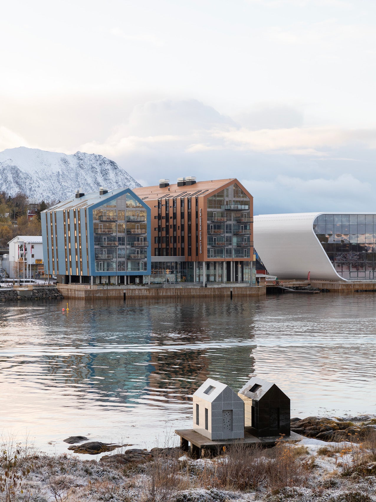 Waterfront modern buildings with glass facades and copper-blue gabled roofs, snow-dusted mountains and reflective harbor