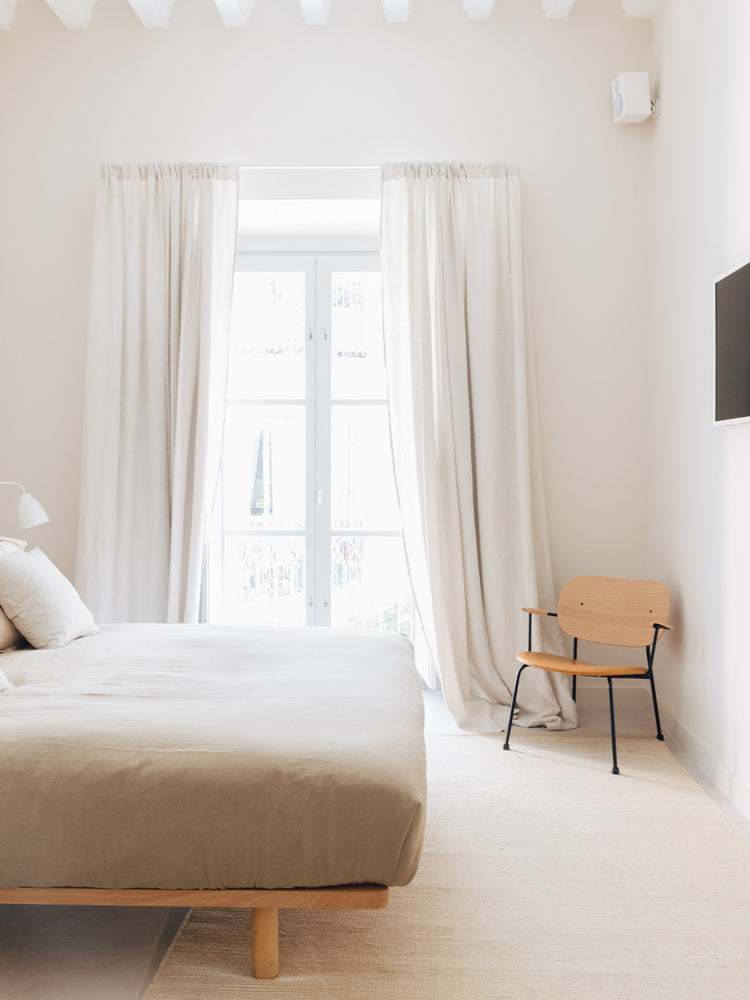 Minimal bedroom with beige linen bed, floor-to-ceiling cream curtains framing a tall window, and a light wooden chair with