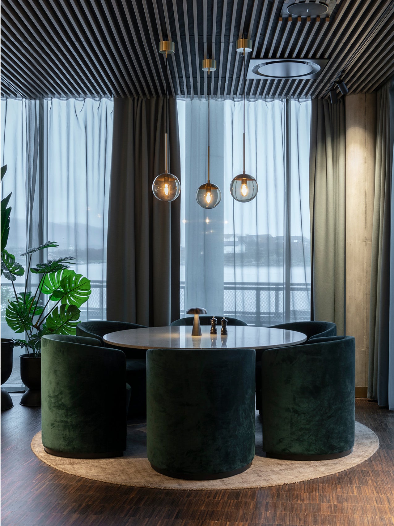 Round marble dining table surrounded by dark green velvet barrel chairs under three brass and glass pendant lights.