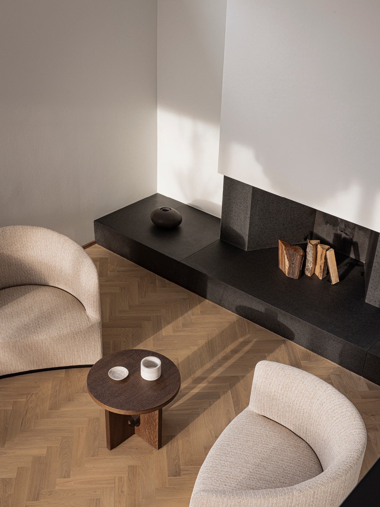 Minimal living room corner with beige curved boucle chairs, dark stone hearth with stacked firewood and round wooden side
