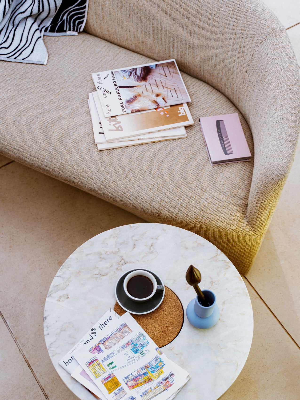 Beige textured curved sofa with stacked magazines and a pink notebook, marble round coffee table with coffee cup and sketches