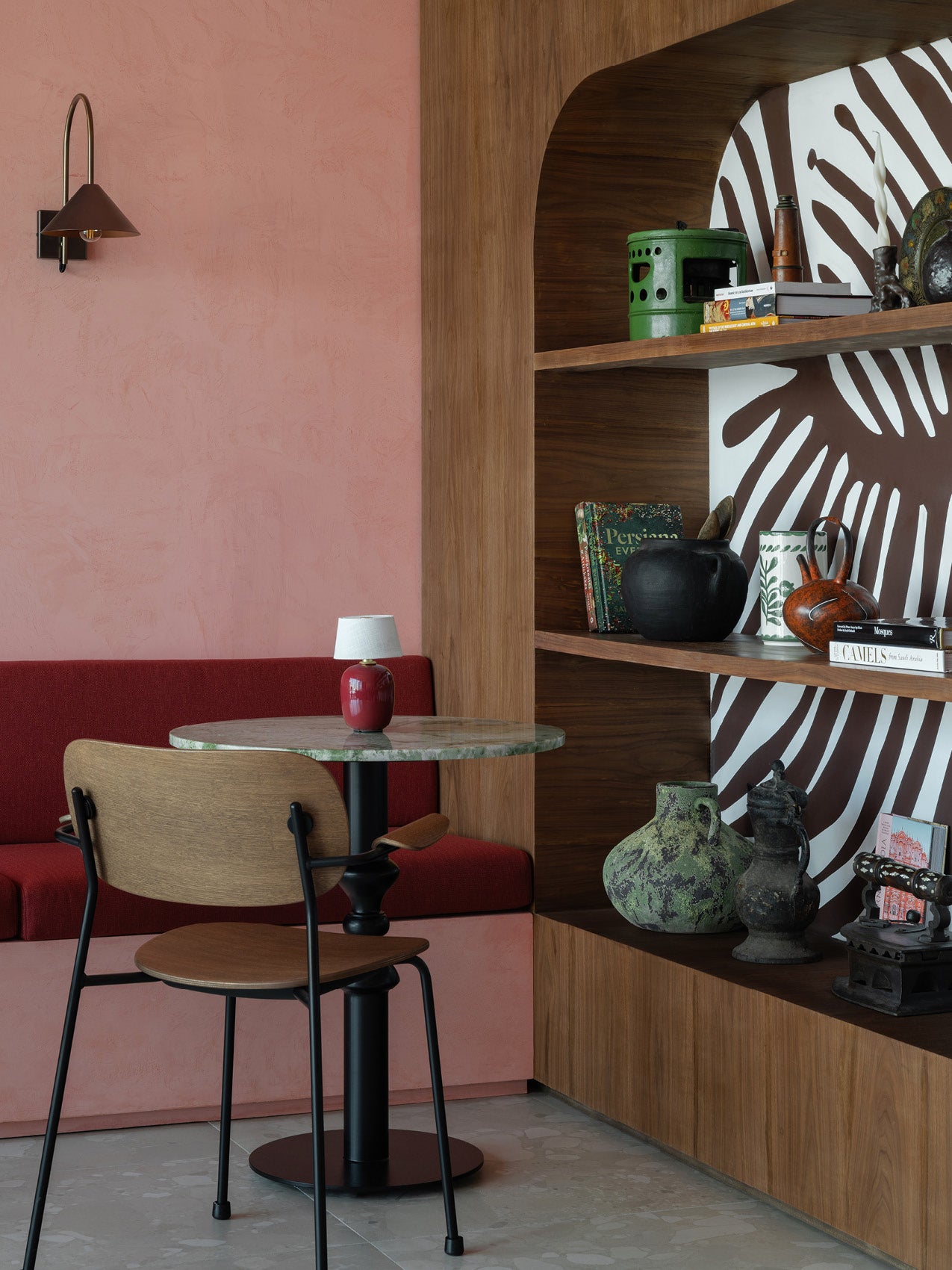 Small round marble-top table with a red ceramic lamp, wooden chair, pink plaster wall and built-in wood shelves with