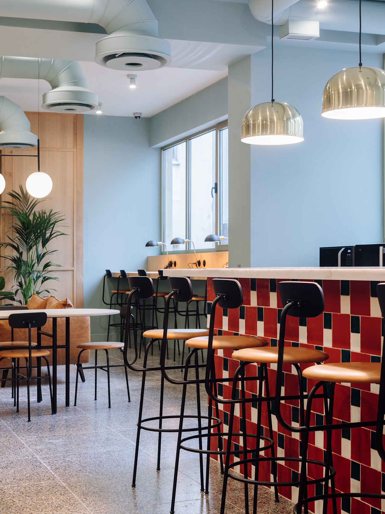 Modern cafe interior with black metal bar stools and tan leather seats at a red-and-white tiled counter, brass pendant