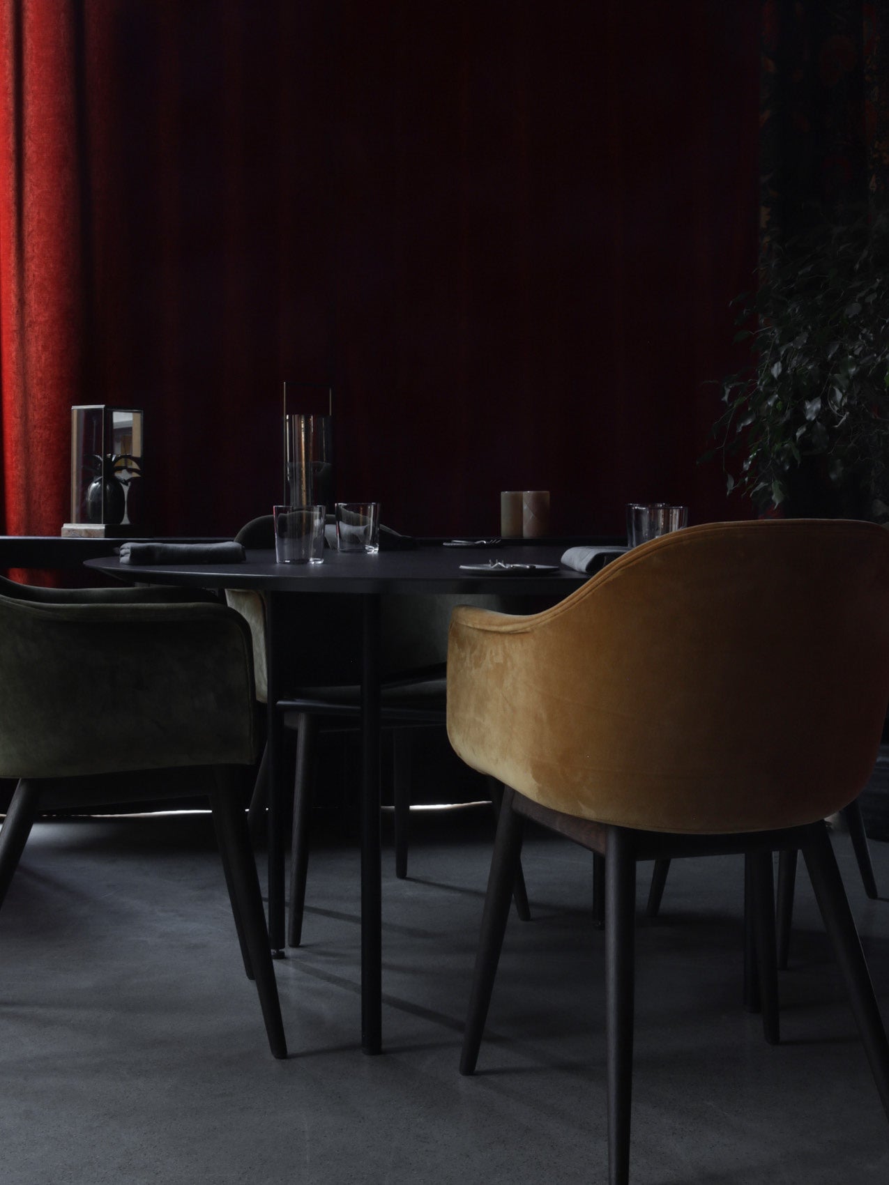 Round black dining table with velvet mustard and olive armchairs, glassware and dark red curtain backdrop