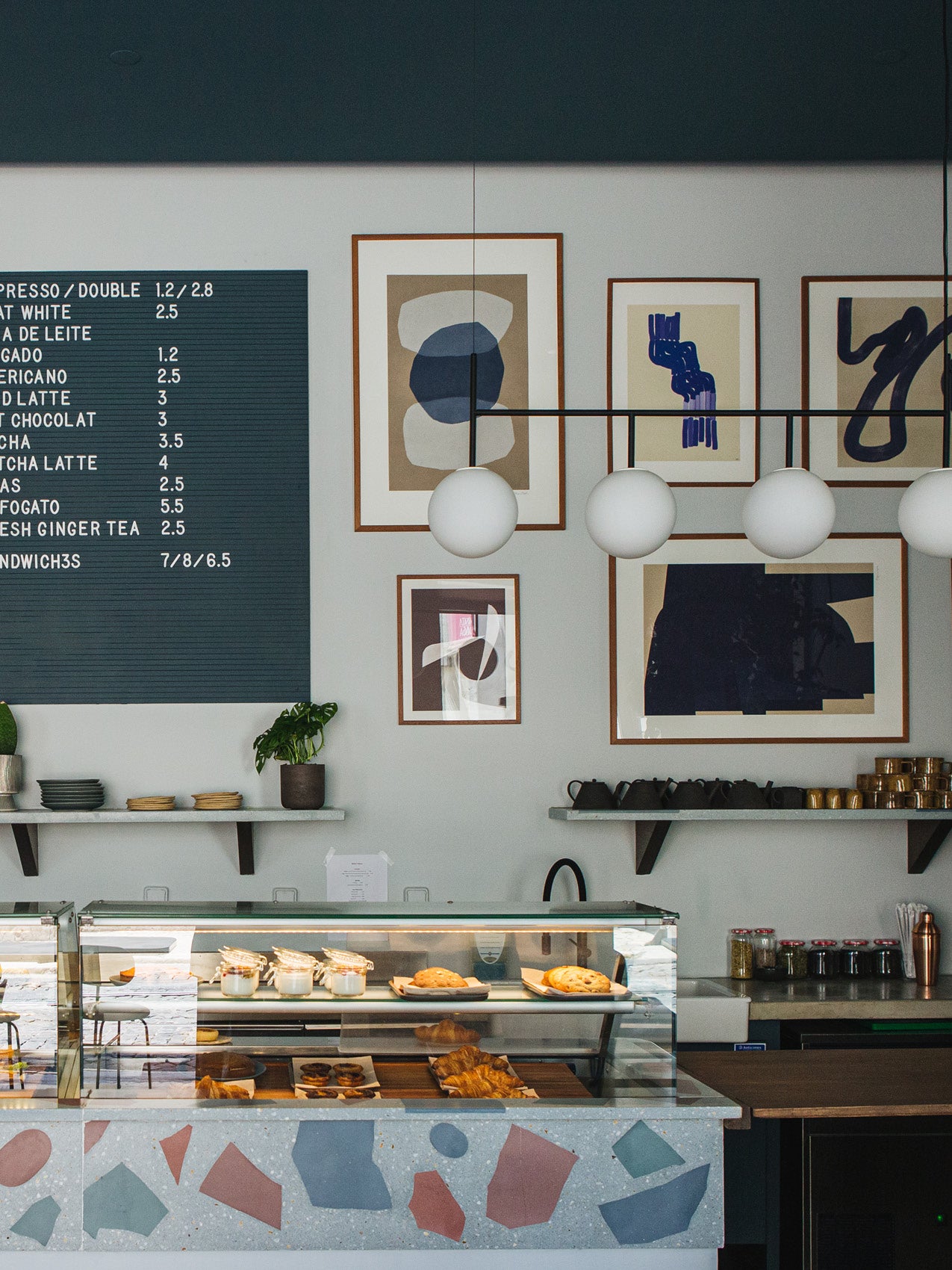 Minimal café counter with terrazzo front, glass pastry case, white globe pendant lights and framed abstract prints on wall