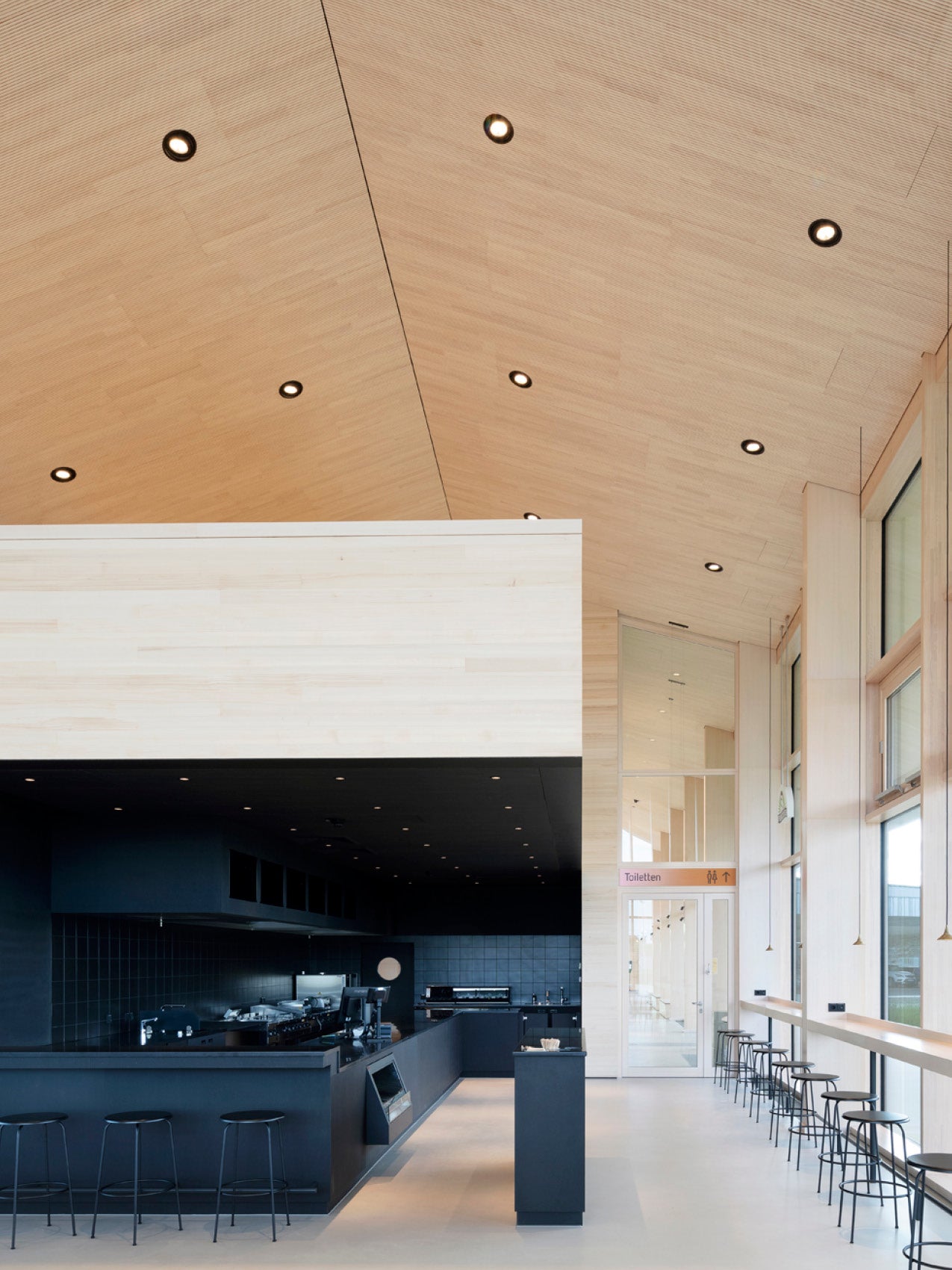 Minimalist cafe interior with light birch wood ceiling and walls, black service counter and row of black metal bar stools by