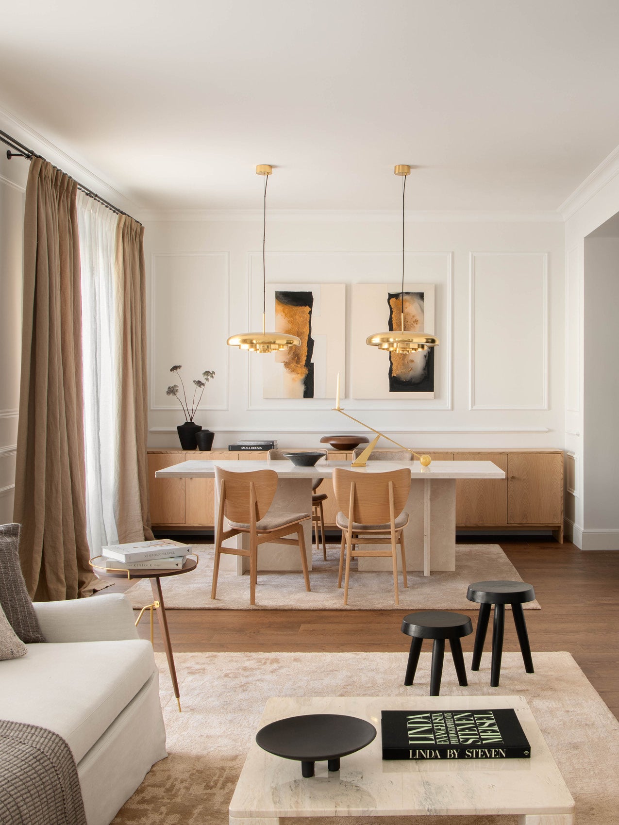 Minimal dining room with marble table, light oak chairs, two brass pendant lights, neutral curtains and abstract wall art
