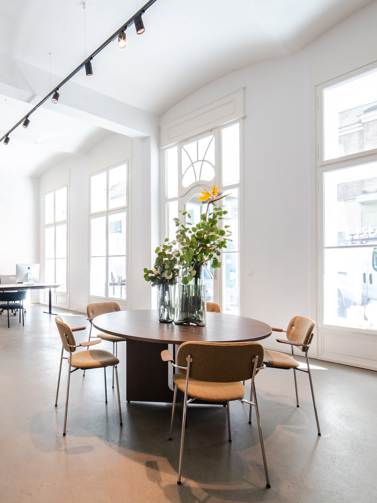 Round dark wood table with glass vases of green foliage and a bird-of-paradise flower, surrounded by tan upholstered chairs
