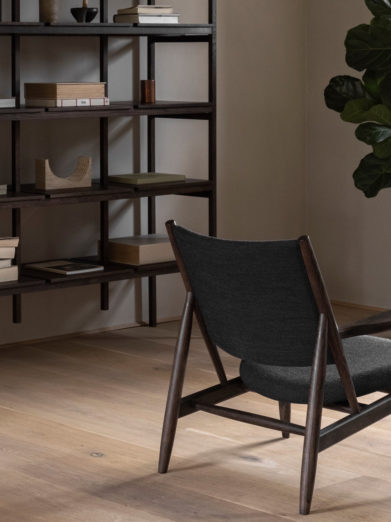 Dark wooden lounge chair with charcoal upholstered seat and curved back, beside a dark oak open shelving unit with books and