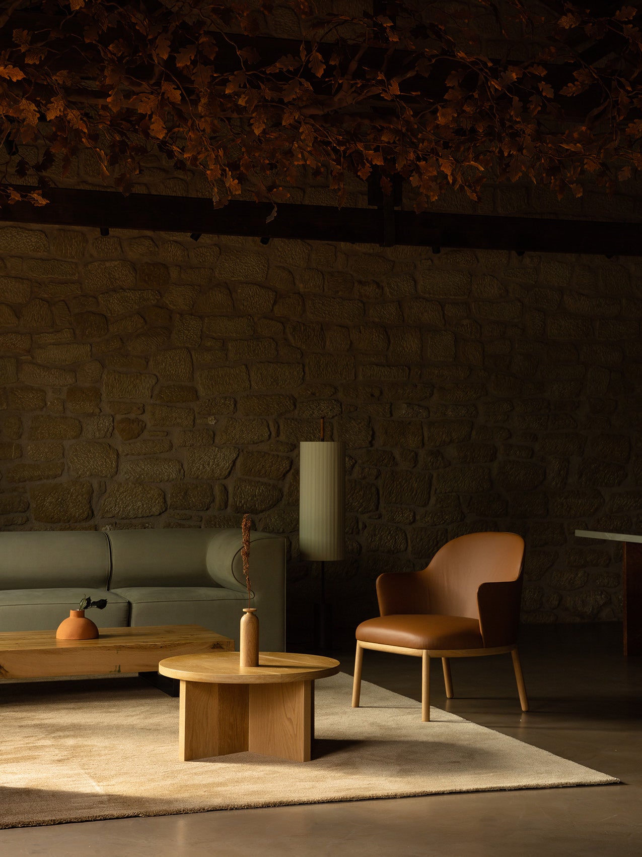 Warm lounge scene with tan leather armchair, olive-green sofa, round oak coffee table and stone wall backdrop