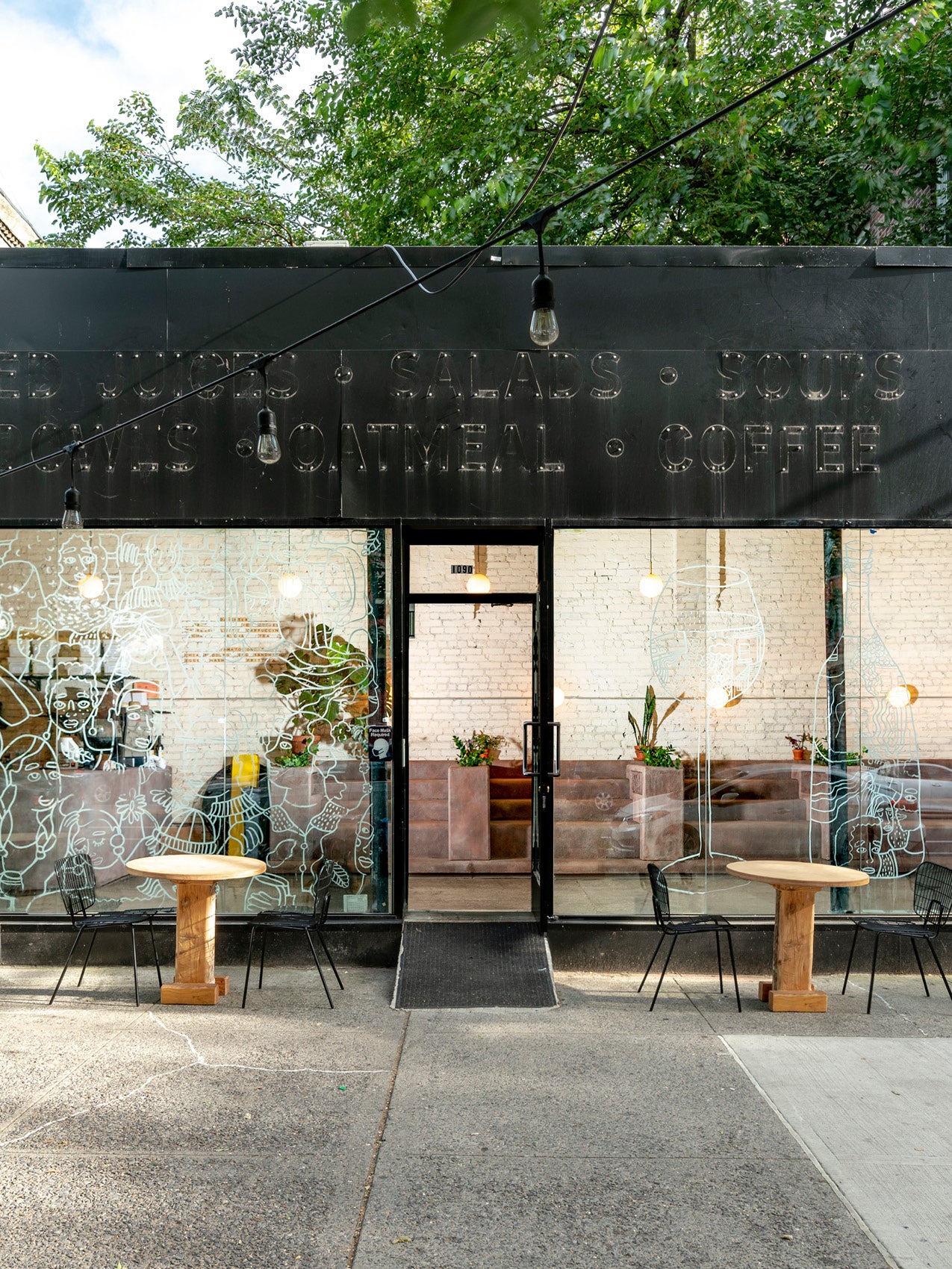 Street-facing café with black metal façade, glass windows with white line art, wooden round tables and black wire chairs.