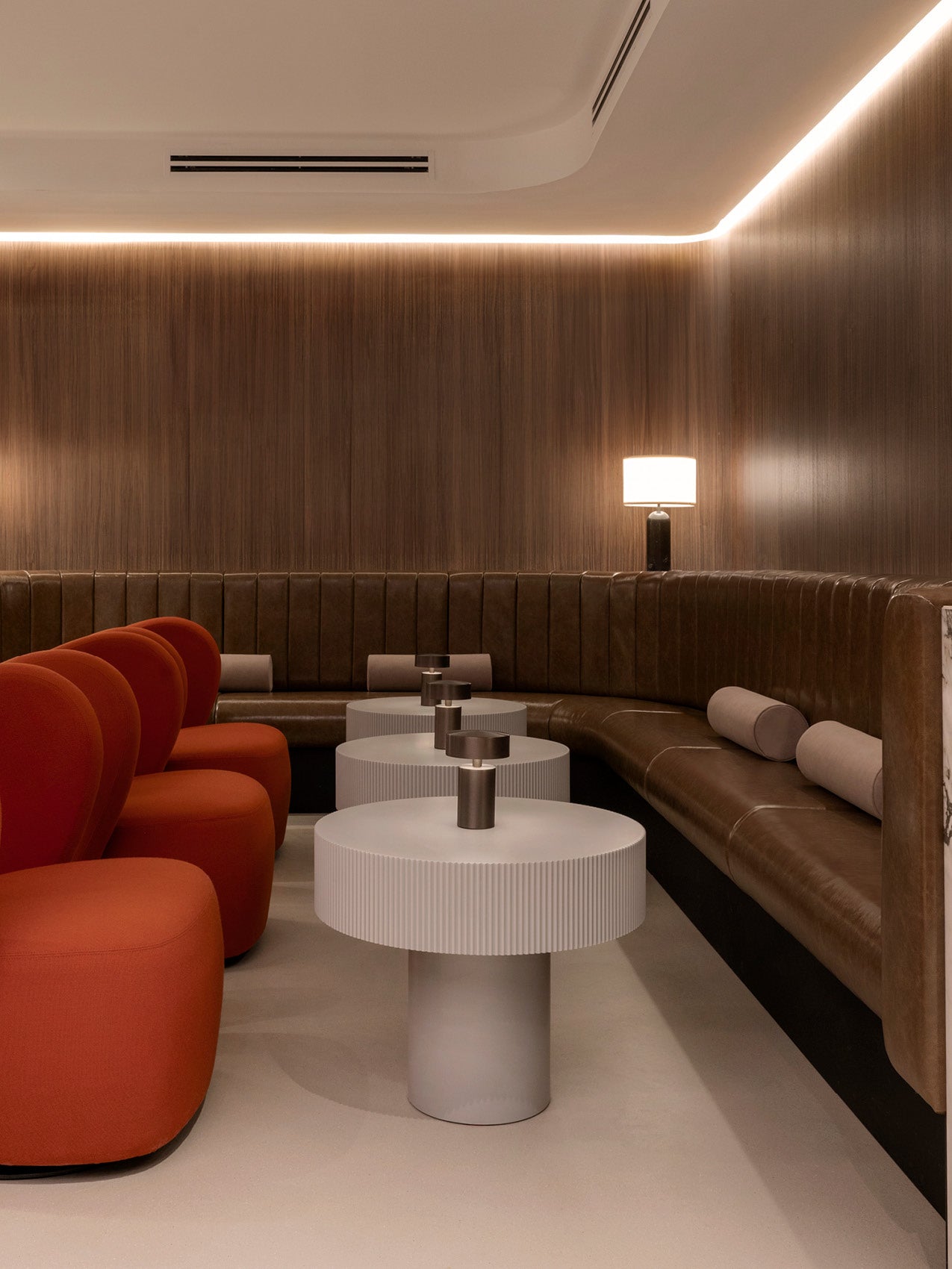Modern lounge with curved brown leather banquette, orange upholstered chairs, ribbed white round side tables and soft