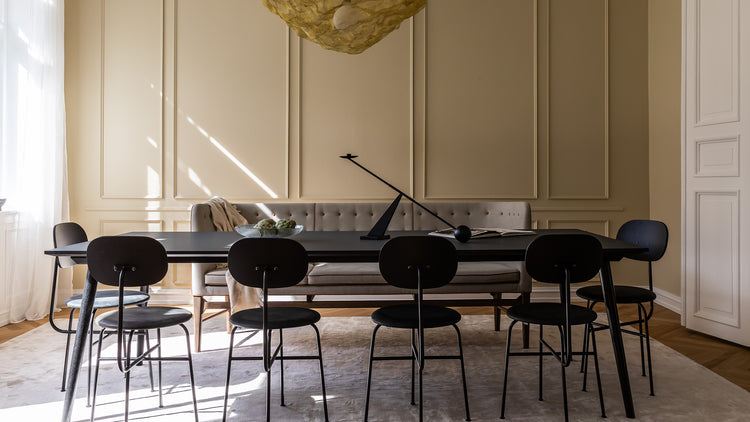 Minimal dining room with long black table, six black metal chairs, beige tufted bench, sculptural lamp and pale yellow wall