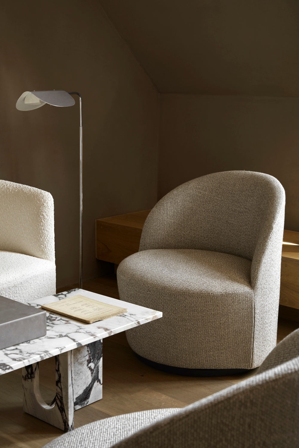 Beige upholstered rounded lounge chair beside a marble coffee table and slender metal floor lamp in warm interior.