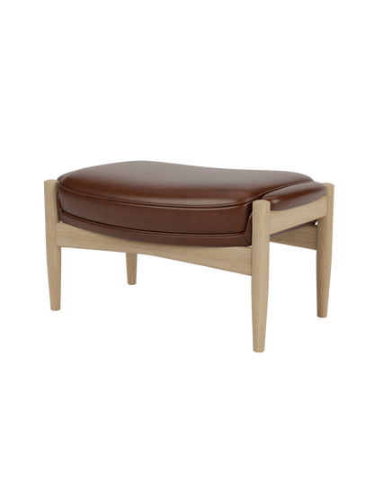 A side-profile view of The Seal Ottoman, showing its plush brown leather cushion and natural oak frame with tapered legs.