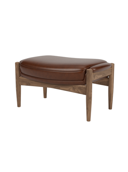 A three-quarter view of The Seal Ottoman, featuring a rich walnut wood frame and a plush, contoured brown leather cushion.