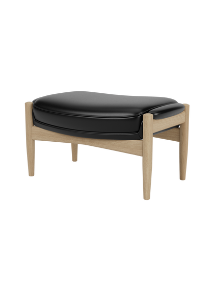 The Seal Ottoman, shown at an angle, features a plush black leather cushion resting on a sculpted natural oak frame.