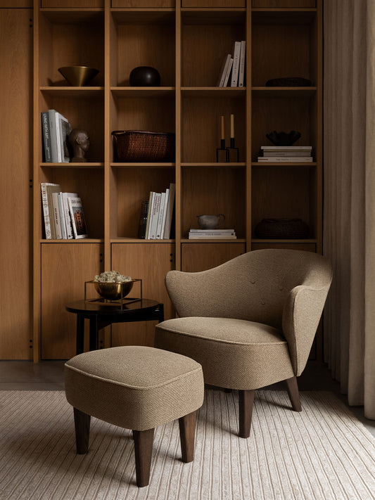 Ingeborg Lounge Chair and Footstool in brown Logan Flint textile with a large brass Kubus Bowl Centerpiece against an oak bookcase.