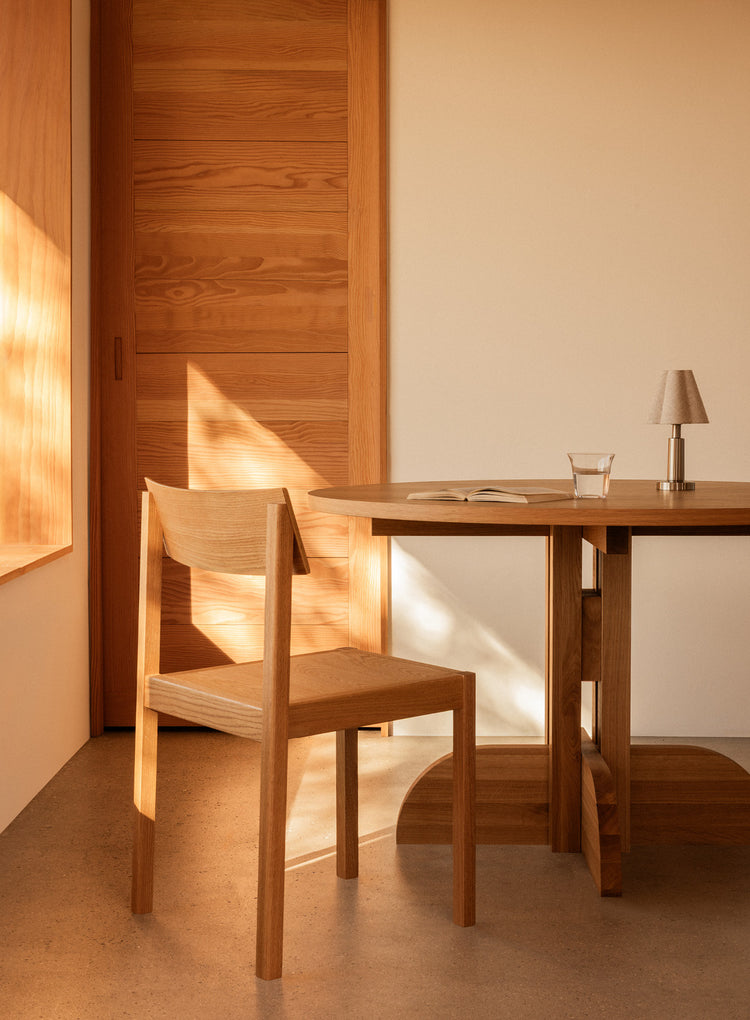 Minimal sunlit dining corner with wooden chair, round table, small lamp and open book