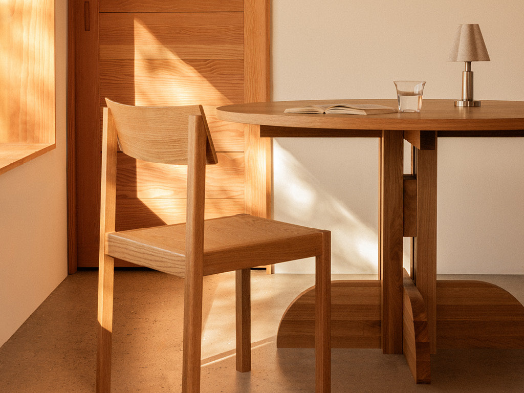 Sunlit minimalist wooden chair and round table with an open book, glass of water and lamp in a warm room