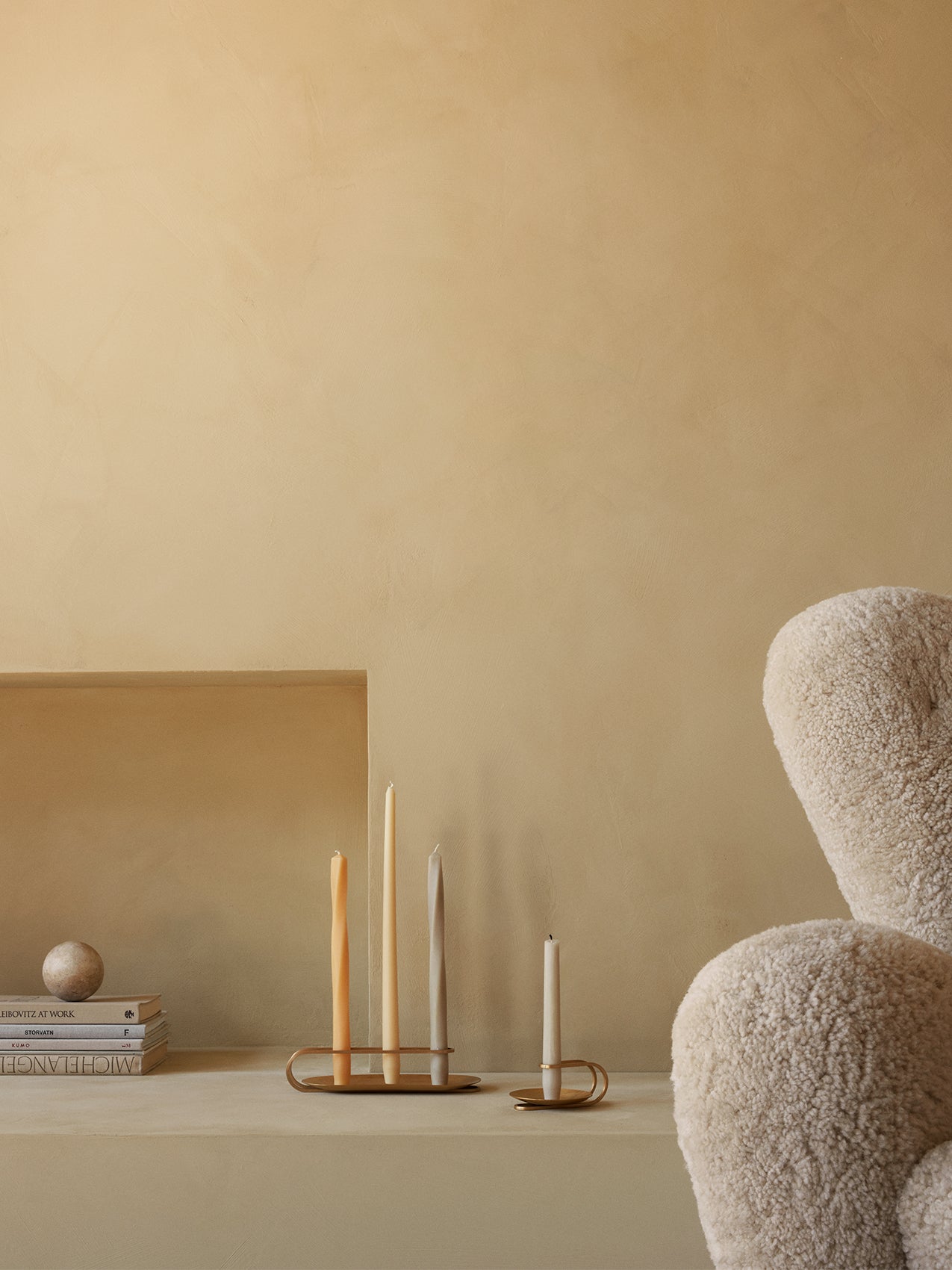 Brass Clip and Table Spire Candle Holders with tapered candles sit on a shelf beside books and a boucle Tired Man armchair.