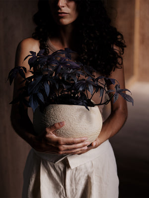 A woman with dark, curly hair holds a large, textured, off-white Plantas Planter filled with a dark-leafed plant.